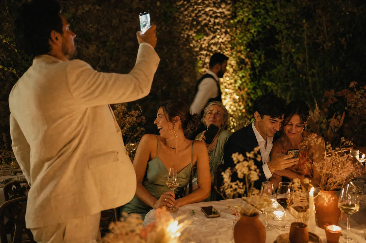Invités à un mariage capturant le moment depuis leur point de vue : un homme debout filme, une femme regarde la scène sans téléphone, un couple partage un même écran
